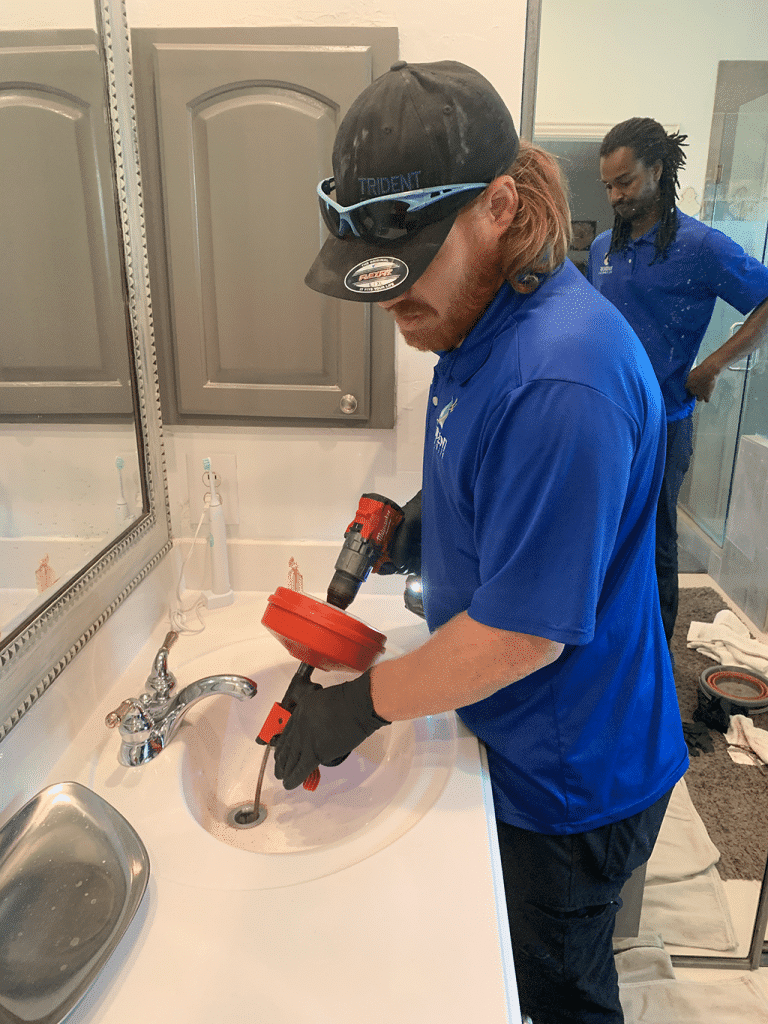 A plumber uses a drain snake to unclog a bathroom sink while another worker stands in the background.