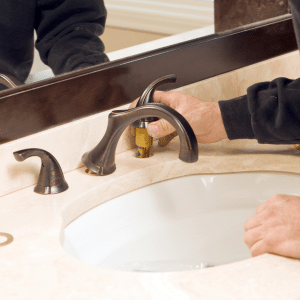 Person installing or repairing a bronze bathroom faucet on a marble countertop with a white sink.