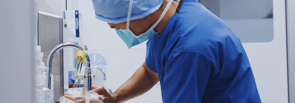 A medical professional in blue scrubs and a mask washes hands at a sink in a clinical setting.