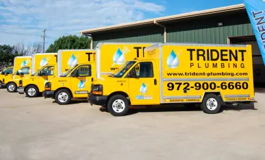 Four yellow Trident Plumbing vans with company logo and contact details are parked outside a green industrial building.