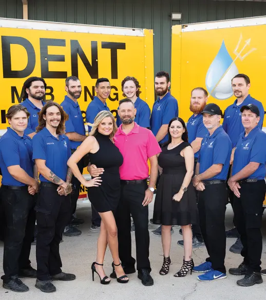A group of thirteen people, mostly men in blue uniforms, pose in front of two yellow service trucks with company logos. Two women in black dresses and a man in a pink shirt stand in front.