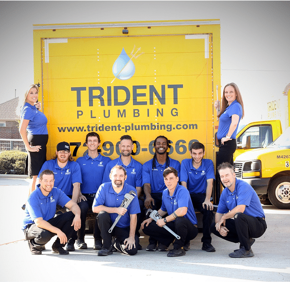 A group of eleven plumbers in blue uniforms pose in front of a yellow Trident Plumbing truck with tools, smiling at the camera.