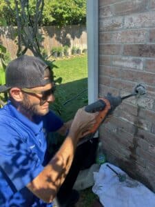 A man wearing sunglasses and a cap uses a power drill to make a hole in a brick wall outside near a garden, possibly preparing for sewer drain pipe repair in Texas.