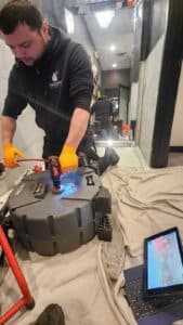 A technician wearing orange gloves uses equipment to inspect or clear a drain as part of a sewer drain pipe repair in Texas, with a tablet displaying footage nearby and tools on a cloth-covered floor.