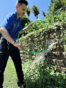 A man in a blue shirt waters plants near a stone wall with a green garden hose on a sunny day, while nearby, signs of recent sewer drain pipe repair in Texas are visible.
