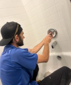 Person in a blue shirt and black cap repairing or installing the faucet handle of a white tiled bathtub, handling bathroom plumbing in Texas.